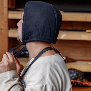 May include: A black quilted hood with tie strings. The hood is made of a dark fabric and features curved stitching patterns. The person is wearing a white shirt and a black strap.