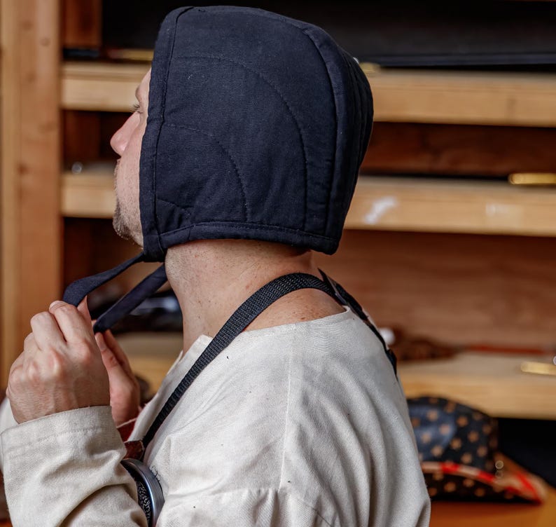 May include: A black quilted hood with tie strings. The hood is made of a dark fabric and features curved stitching patterns. The person is wearing a white shirt and a black strap.