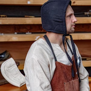 May include: A black quilted hood with ties, worn by a person. The hood is part of a historical costume, paired with a white shirt and a brown leather apron. The background features wooden shelves with various items.