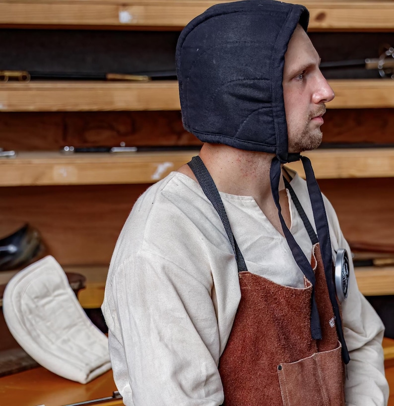 May include: A black quilted hood with ties, worn by a person. The hood is part of a historical costume, paired with a white shirt and a brown leather apron. The background features wooden shelves with various items.