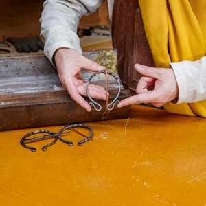 May include: A person holding a handcrafted metal bracelet with a copper accent. The bracelet has a unique, open design with curled ends. Other metalwork pieces are visible on a yellow surface.