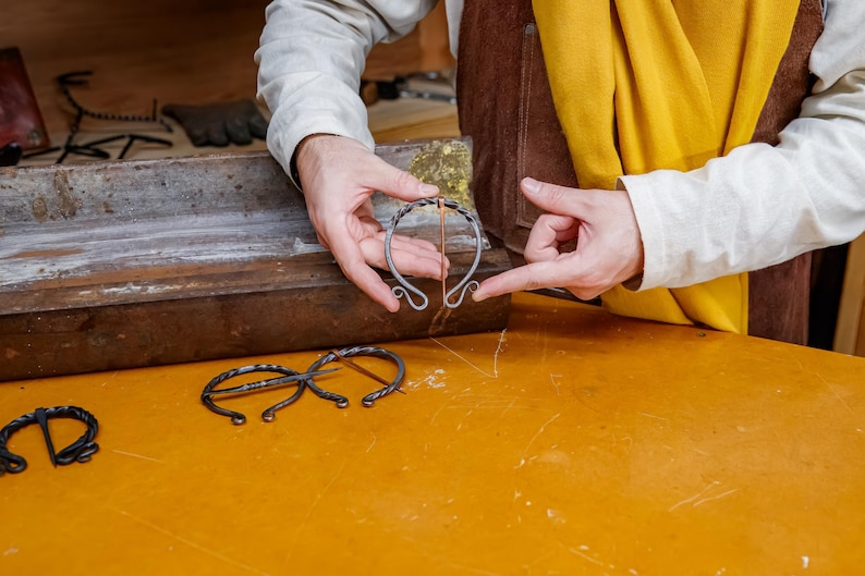 May include: A person holding a handcrafted metal bracelet with a copper accent. The bracelet has a unique, open design with curled ends. Other metalwork pieces are visible on a yellow surface.