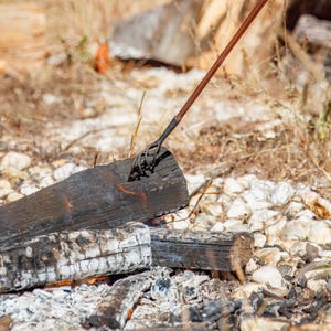 May include: A metal fire poker with a spiral end is inserted into a charred log. The poker has a wooden handle. The scene includes burning wood, ash, and dry grass, suggesting a campfire or outdoor fire pit.