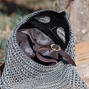 May include: A medieval chainmail coif with a leather helmet and chin strap. The coif is made of small, interlocking metal rings. The helmet is brown with a leather chin strap and a brass buckle. The coif is draped over a wooden surface.
