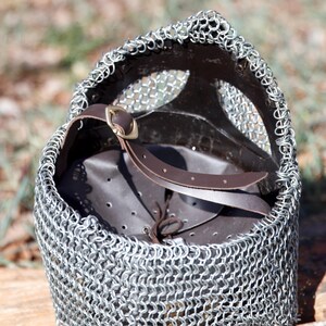 May include: A close-up of a medieval chainmail coif. The coif is made of small, interlocking metal rings and features a brown leather interior and a brown leather strap with a brass buckle. The coif is designed to protect the head and neck.