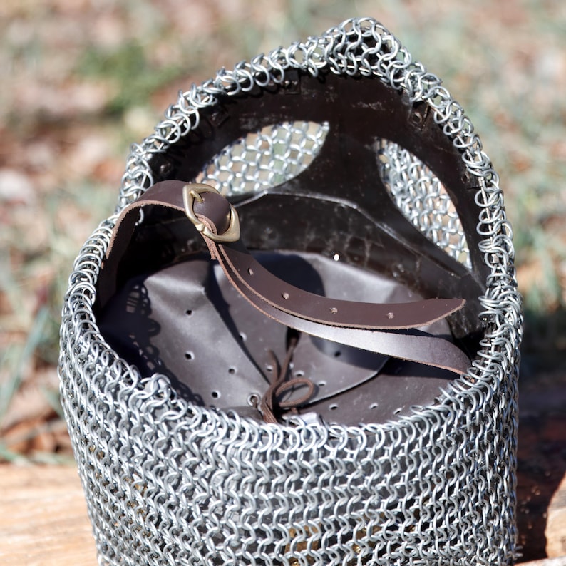 May include: A close-up of a medieval chainmail coif. The coif is made of small, interlocking metal rings and features a brown leather interior and a brown leather strap with a brass buckle. The coif is designed to protect the head and neck.