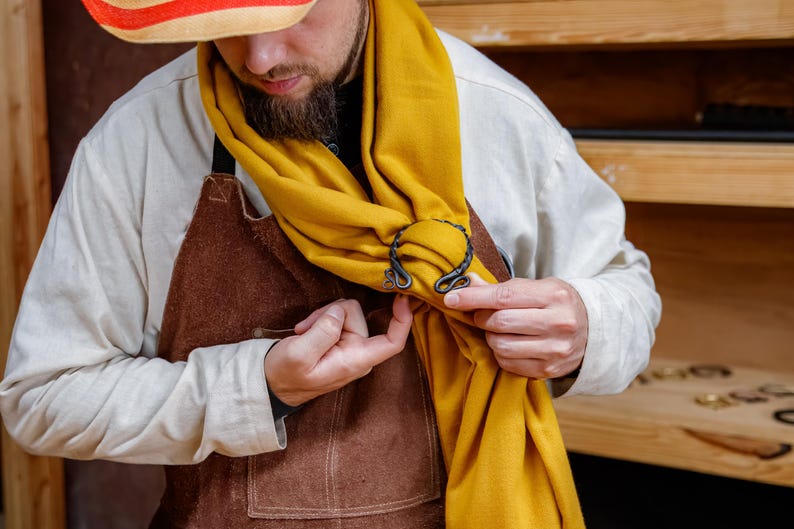 May include: A mustard yellow scarf is held in place by a decorative, dark metal scarf ring. The scarf ring has a unique, swirling design. The person in the image is wearing a white shirt, brown apron, and a straw hat with red and orange stripes.