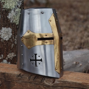 May include: A silver medieval helmet with brass detailing. The helmet has a cross design and is riveted. Brass elements adorn the front and sides, enhancing the historical armour's appearance. A piece of historical headwear.