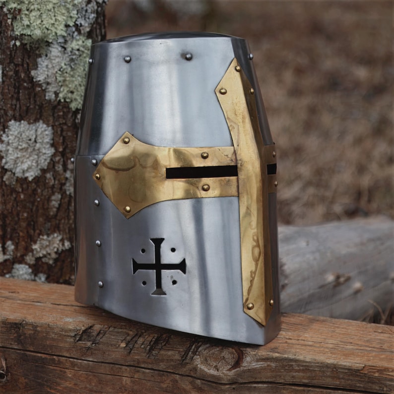 May include: A silver medieval helmet with brass detailing. The helmet has a cross design and is riveted. Brass elements adorn the front and sides, enhancing the historical armour's appearance. A piece of historical headwear.