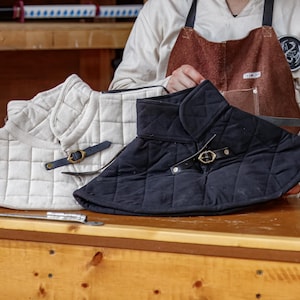 May include: Two quilted neck protectors, one cream and one black, with leather straps and brass buckles. The protectors are displayed on a wooden surface, showcasing their diamond-stitched pattern and protective design.