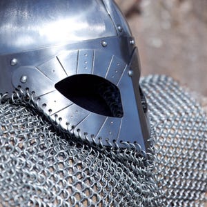 May include: A close-up of a silver Viking helmet with a riveted design and eye opening. The helmet is partially covered with chainmail. The helmet is made of metal and has a polished finish.