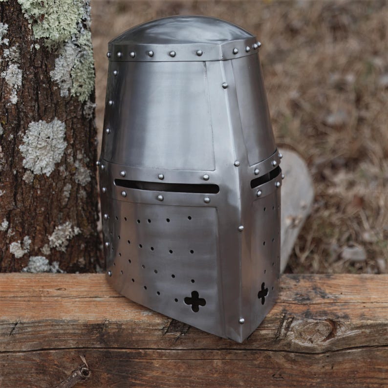 May include: A silver-coloured medieval crusader helmet. The helmet features a flat top, a rectangular face opening, and small rivet details. The metal helmet is displayed on a wooden surface. The helmet's design is reminiscent of historical armour.
