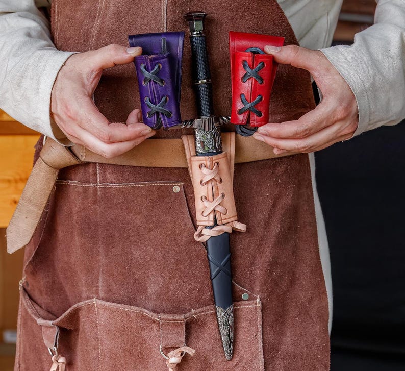 May include: A medieval dagger with a black handle and silver detailing, presented in a tan leather sheath. Two small leather pouches, purple and red, are held nearby. The dagger and sheaths are displayed against a brown leather apron.