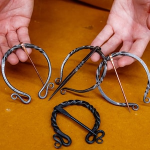 May include: Hand-forged metal brooches in various designs. The brooches feature a circular or U-shaped frame with a pin. The metal has a dark, textured appearance, with some pins in a copper color. The brooches are displayed on a yellow-brown surface.