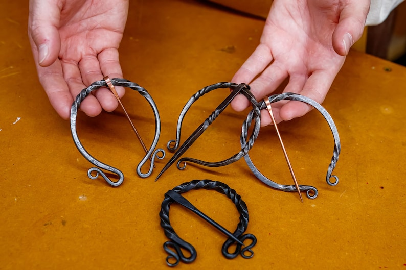 May include: Hand-forged metal brooches in various designs. The brooches feature a circular or U-shaped frame with a pin. The metal has a dark, textured appearance, with some pins in a copper color. The brooches are displayed on a yellow-brown surface.