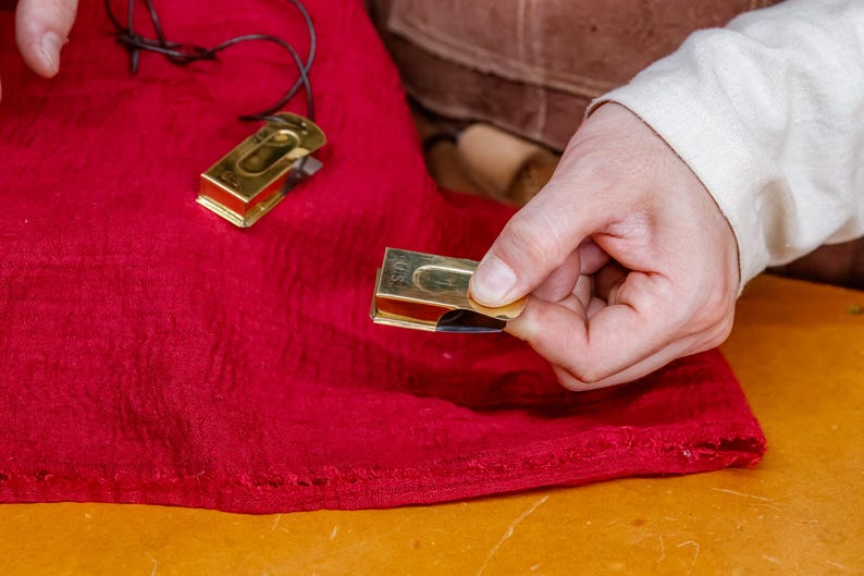 Pu&ograve; includere: Primo piano di due oggetti metallici rettangolari color oro con un inserto nero, uno tenuto in mano. Gli oggetti sono su un tessuto rosso. Un oggetto ha un cordino nero attaccato. Gli oggetti sembrano piccoli strumenti.