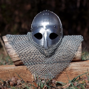 May include: A silver medieval helmet with a riveted design and a long nose guard. The helmet is paired with a chainmail coif, displayed on a wooden surface. The background is blurred, suggesting an outdoor setting.
