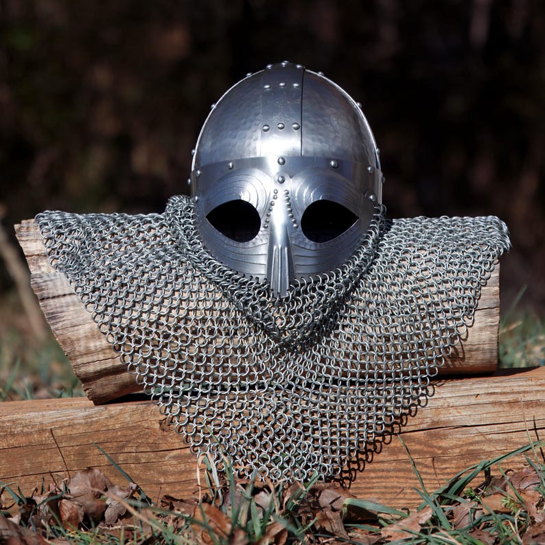 May include: A silver medieval helmet with a riveted design and a long nose guard. The helmet is paired with a chainmail coif, displayed on a wooden surface. The background is blurred, suggesting an outdoor setting.