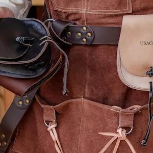 Puede incluir: Un delantal de cuero marr&oacute;n con un cintur&oacute;n de cuero marr&oacute;n y dos bolsas. La bolsa m&aacute;s grande es de color beige y tiene el texto "USACraftmen" impreso en ella. La bolsa m&aacute;s peque&ntilde;a es marr&oacute;n y tiene una solapa.