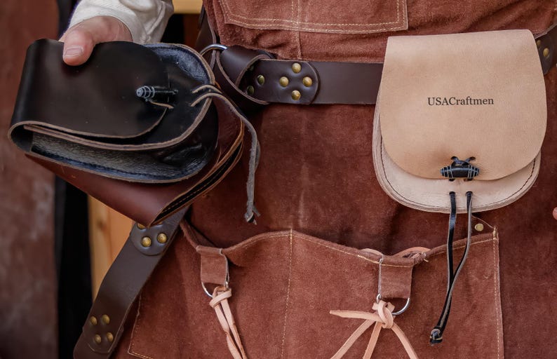 Puede incluir: Un delantal de cuero marr&oacute;n con un cintur&oacute;n de cuero marr&oacute;n y dos bolsas. La bolsa m&aacute;s grande es de color beige y tiene el texto "USACraftmen" impreso en ella. La bolsa m&aacute;s peque&ntilde;a es marr&oacute;n y tiene una solapa.