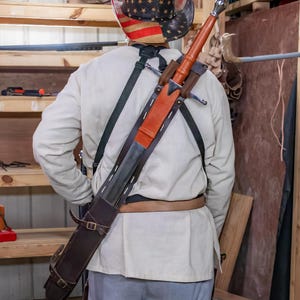 May include: A person wearing a white shirt and a cowboy hat with an American flag design is wearing a brown leather sword scabbard with a silver sword strapped across their back.