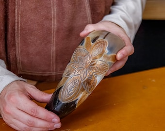 May include: A carved horn drinking vessel with a floral design. The horn is a mix of brown and tan colors, with a dark brown base. The floral pattern is etched into the horn's surface, creating a detailed, textured appearance.