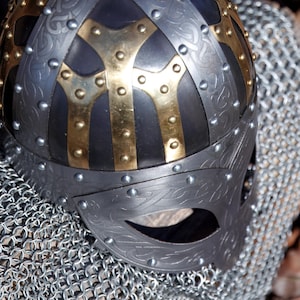 May include: A close-up of a Viking helmet. The helmet is dark grey with gold accents and rivets. It features decorative patterns and a face guard. The helmet rests on a chainmail surface.