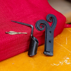 May include: An assortment of metal hardware pieces, including a black hinge with decorative scrollwork, a black metal tool, and a small, ornate brass piece. The items are arranged on a red fabric and a yellow surface.