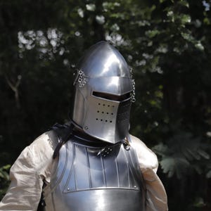 Pu&ograve; includere: Un elmo da cavaliere in acciaio lucido con sommit&agrave; appuntita, una fessura orizzontale per gli occhi e una visiera perforata. L'elmo presenta piccole catene metalliche fissate ai lati ed &egrave; indossato sopra una corazza e una camicia chiara.