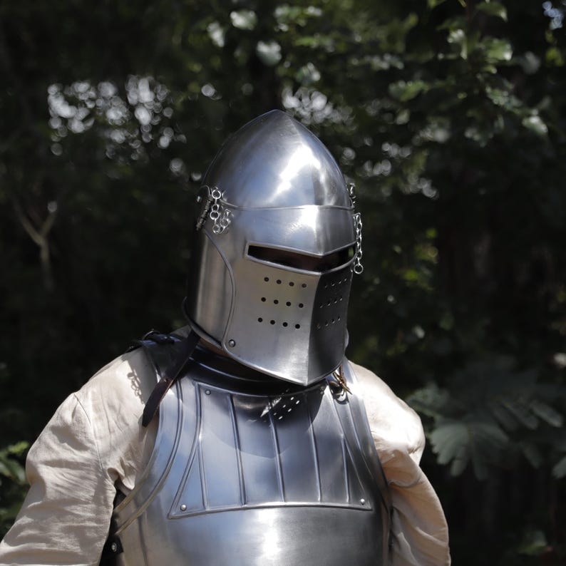 Pu&ograve; includere: Un elmo da cavaliere in acciaio lucido con sommit&agrave; appuntita, una fessura orizzontale per gli occhi e una visiera perforata. L'elmo presenta piccole catene metalliche fissate ai lati ed &egrave; indossato sopra una corazza e una camicia chiara.