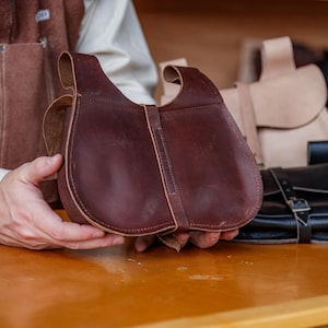 Op de afbeelding: Een bruine leren tas met een klep sluiting en een gesp. De tas wordt vastgehouden door een hand.