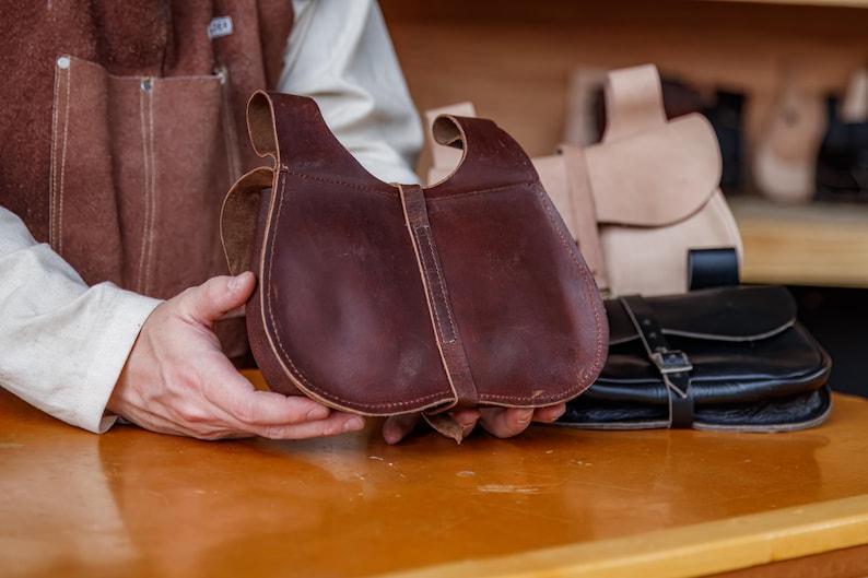 Op de afbeelding: Een bruine leren tas met een klep sluiting en een gesp. De tas wordt vastgehouden door een hand.
