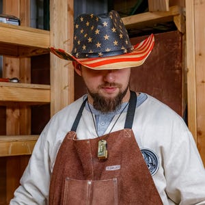 Pu&ograve; includere: Un cappello da cowboy con un design patriottico. Il cappello ha una corona nera con stelle dorate e un bordo a righe rosse e bianche. Viene indossato con un grembiule di pelle marrone e una camicia bianca.