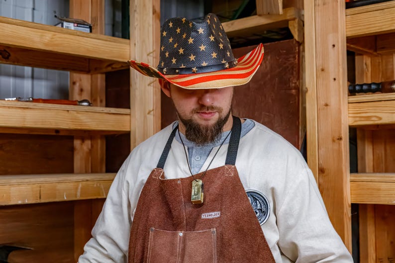 Pu&ograve; includere: Un cappello da cowboy con un design patriottico. Il cappello ha una corona nera con stelle dorate e un bordo a righe rosse e bianche. Viene indossato con un grembiule di pelle marrone e una camicia bianca.