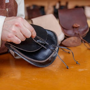 Puede incluir: Una bolsa de cuero negro con cierre de solapa y cord&oacute;n de ajuste. La bolsa est&aacute; siendo sostenida por la mano de una persona. La bolsa est&aacute; sobre una mesa de madera con otras bolsas de cuero en el fondo.