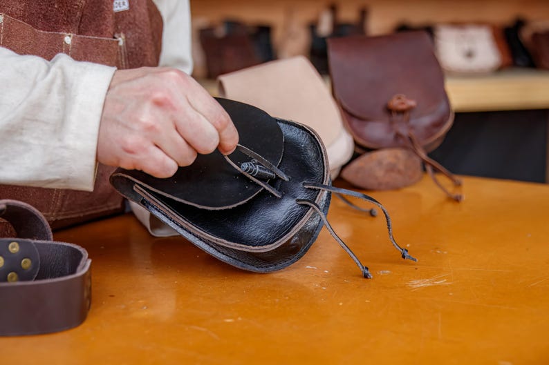 Puede incluir: Una bolsa de cuero negro con cierre de solapa y cord&oacute;n de ajuste. La bolsa est&aacute; siendo sostenida por la mano de una persona. La bolsa est&aacute; sobre una mesa de madera con otras bolsas de cuero en el fondo.
