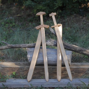 May include: Two light-colored wooden swords featuring carved geometric patterns on the hilts and crossguards. The blades are smooth and straight, with one sword positioned slightly behind the other, showcasing detailed craftsmanship on the handles.