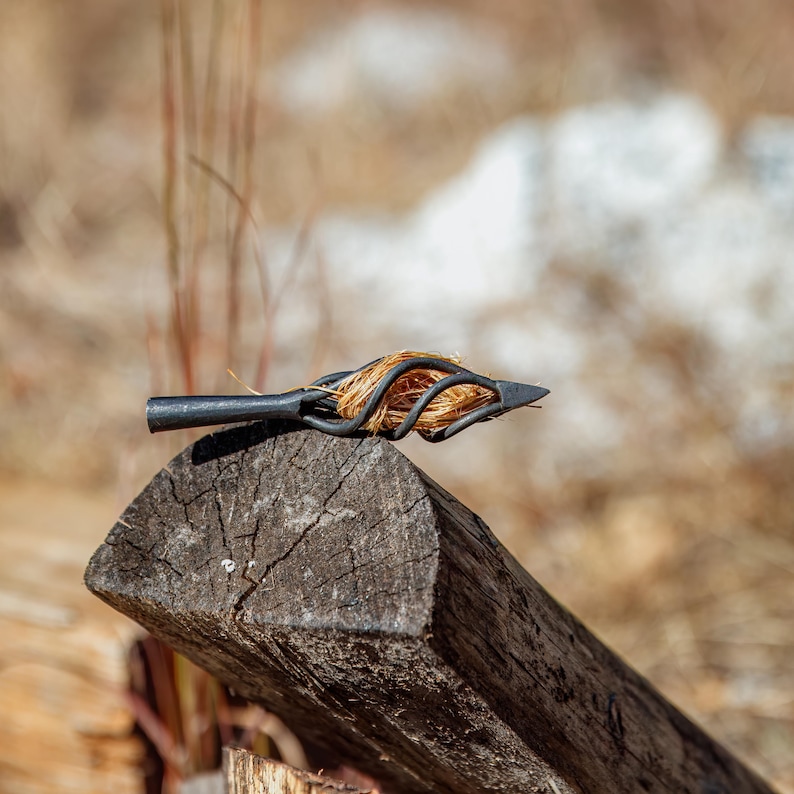 May include: A handcrafted, black metal spearhead with a woven, golden-colored element, resting on a weathered wooden log. The spearhead's design features a pointed tip and a decorative, looped structure.