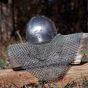 May include: A silver medieval helmet with a riveted design, displayed with a chainmail coif. The helmet sits on a wooden surface. The background is a blurred natural setting, evoking a historical theme.