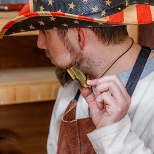 Pu&ograve; includere: Un cappello da cowboy di paglia con un design patriottico raffigurante la bandiera americana. Il cappello ha una fascia in pelle nera ed &egrave; decorato con stelle dorate. La persona tiene in mano un piccolo oggetto metallico dorato.