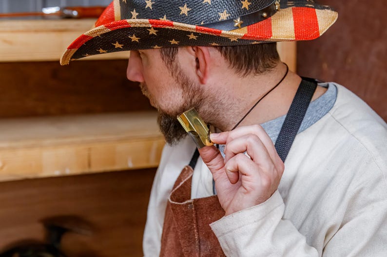 Pu&ograve; includere: Un cappello da cowboy di paglia con un design patriottico raffigurante la bandiera americana. Il cappello ha una fascia in pelle nera ed &egrave; decorato con stelle dorate. La persona tiene in mano un piccolo oggetto metallico dorato.