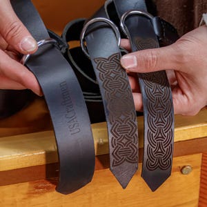 May include: Three black leather belts with different engraved designs. The belt on the left has the text "USACraftmen" engraved on it. The other two belts have intricate Celtic knot designs.