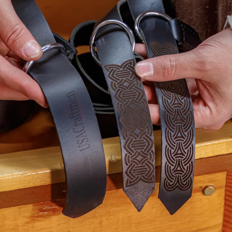 May include: Three black leather belts with different engraved designs. The belt on the left has the text "USACraftmen" engraved on it. The other two belts have intricate Celtic knot designs.
