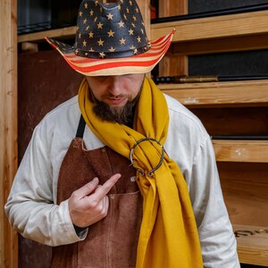 May include: A person wearing a straw cowboy hat with a black crown and gold stars, and red and white stripes on the brim. They are wearing a yellow scarf secured with a metal ring, a brown leather apron, and a white shirt.