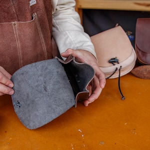 Puede incluir: Una persona sostiene una bolsa de cuero marr&oacute;n abierta, mostrando el forro de tela gris oscuro. La bolsa tiene una solapa y un cierre de hebilla. Hay otras dos bolsas de cuero en el fondo, una es marr&oacute;n claro y la otra es marr&oacute;n oscuro.