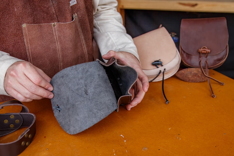 Puede incluir: Una persona sostiene una bolsa de cuero marr&oacute;n abierta, mostrando el forro de tela gris oscuro. La bolsa tiene una solapa y un cierre de hebilla. Hay otras dos bolsas de cuero en el fondo, una es marr&oacute;n claro y la otra es marr&oacute;n oscuro.