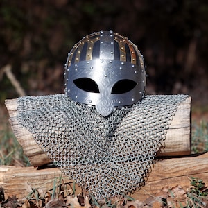 May include: A silver Viking helmet with gold and silver detailing, including a face guard with eye openings. The helmet sits on a wooden log, with a chainmail coif below. The background is blurred, with natural foliage.
