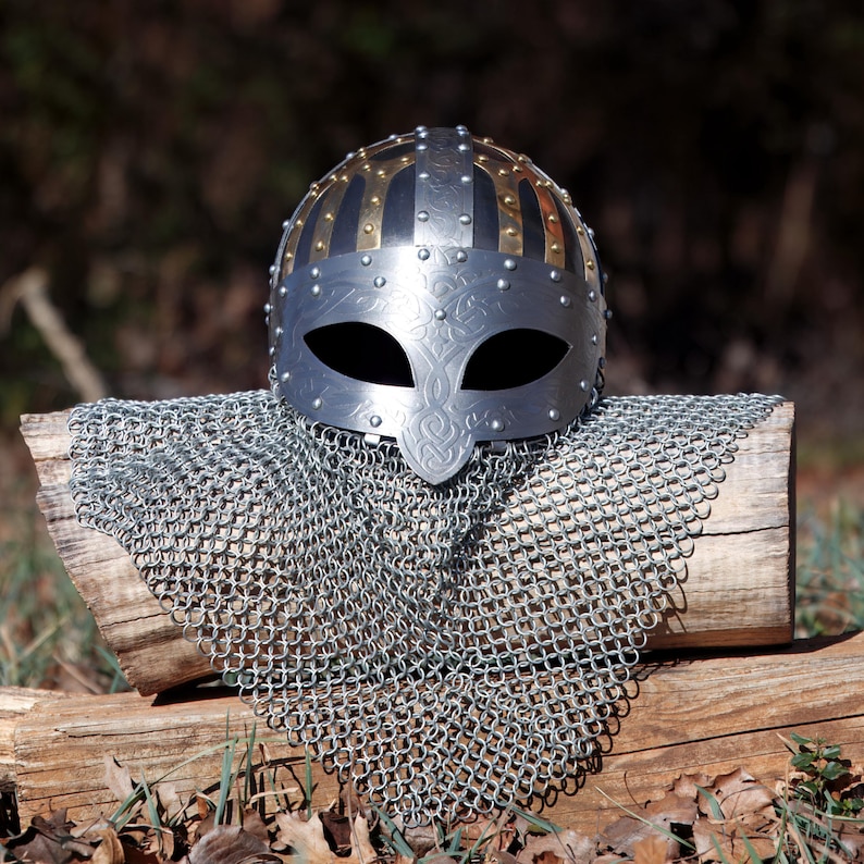 May include: A silver Viking helmet with gold and silver detailing, including a face guard with eye openings. The helmet sits on a wooden log, with a chainmail coif below. The background is blurred, with natural foliage.
