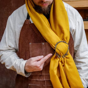 May include: A mustard yellow scarf is held in place by an ornate, antique-style metal brooch. The brooch has a decorative, swirling design. The person in the image is wearing a brown leather apron and a white shirt.