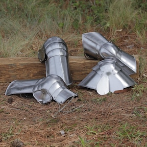 May include: Four pieces of medieval armor are displayed on a bed of pine needles and a wooden beam. The armor includes two arm guards, a shoulder guard, and a knee guard, all made of shiny, silver-colored metal.